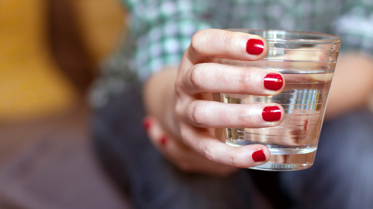 Eine Frau mit lackierten Fingernägeln hält ein Glas Trinkwasser in der Hand.