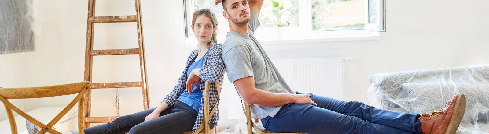 Junges Paar sitzt erschöpft und müde beim Einzug in eine neue Wohnung auf zwei Stühlen, die auf ausgefalteten Pappkartons stehen. Hinten an der Wand ist eine Leiter angelehnt. Das Sofa ist noch in Folie eingepackt.
