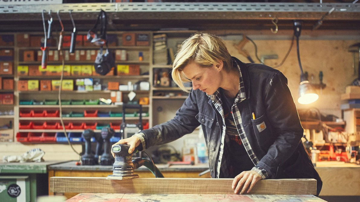 Eine Frau schleift mit einer Maschine ein Stück Holz.