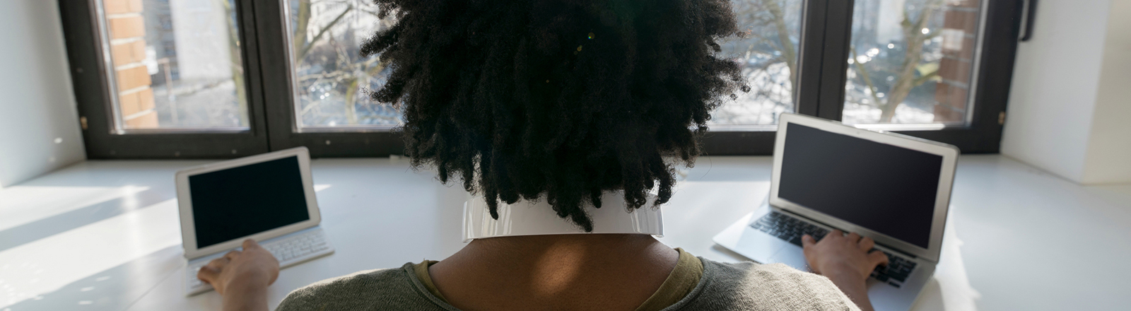 Ein Mann arbeitet zu Hause vor einem Panoramafenster, auf dem Tisch vor ihm steht links ein Tablett und rechts ein Laptop; der Mann ist von hinten zu sehen