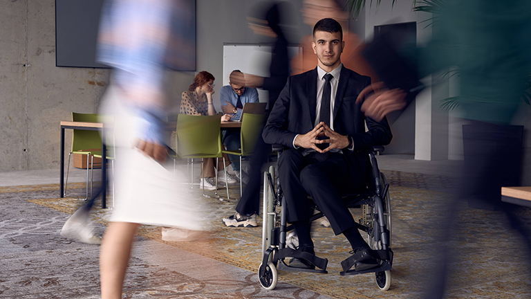 Ein Mann sitzt in einem Rollstuhl, um ihn herum laufen Menschen. Der Raum wirkt wie ein Arbeitsplatz.