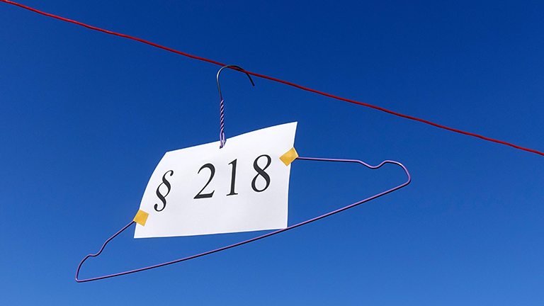 Vor blauem Himmel hängt ein Drahtbügel an einem Seil, daran hängt ein Schild auf dem Paragraf 218 steht.