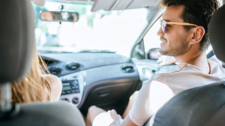 Ein Mann sitzt auf dem Beifahrersitz eines Autos, er blickt zur Fahrerin. Das Foto zeigt beide von hinten.