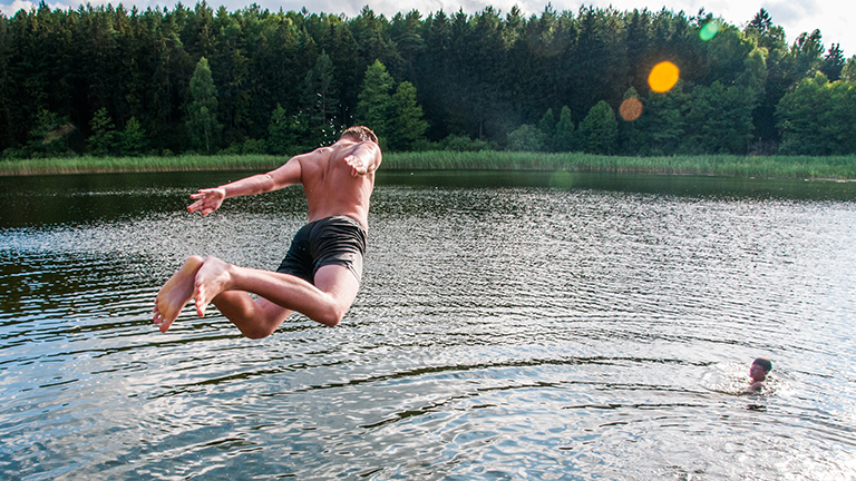 Ein junger Mann springt in einen See, ein andere ist schon im Wasser. Sommerliche Atmosphäre.