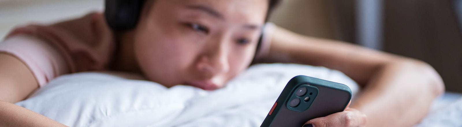 Eine Frau liegt auf dem Bauch in einem Bett und schaut auf ihr Smartphone; sie wirkt traurig.