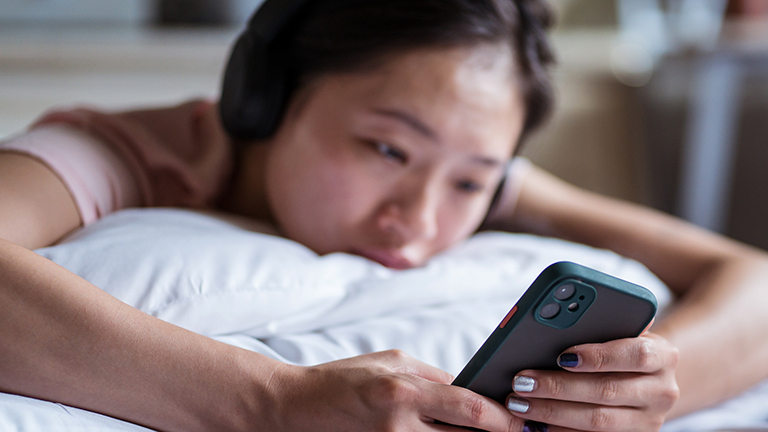Eine Frau liegt auf dem Bauch in einem Bett und schaut auf ihr Smartphone; sie wirkt traurig.