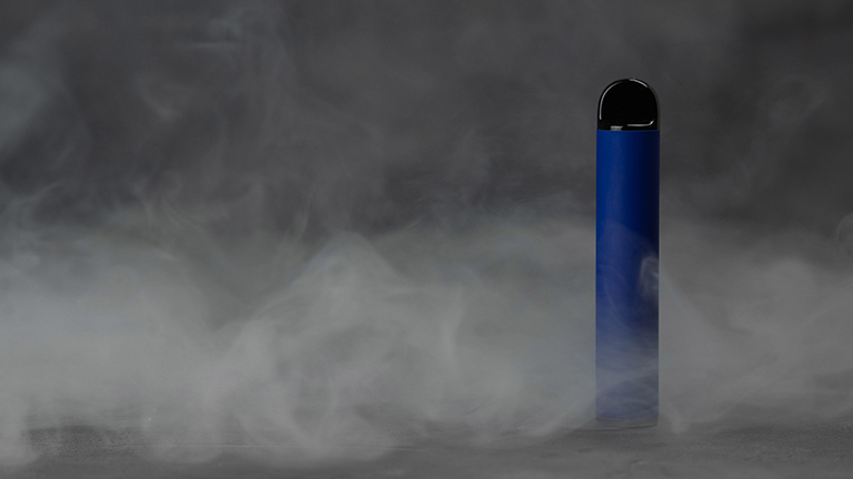 Ein blauer Vaper steht vor dunklem Hintergrund und ist von Rauch umhüllt.