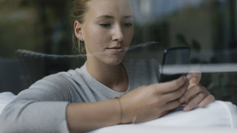 Eine junge Frau sitzt auf einem Sofa, hinter einer Fensterscheibe. Sie hält mit beiden Händen ein Smartphone und blickt darauf.