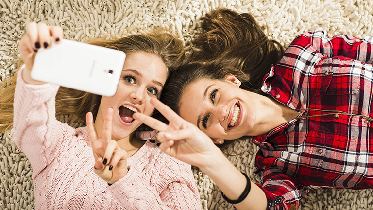 Zwei junge Frauen liegen nebeneinander auf einem Teppich. Eine hält ein Smartphone in die Höhe und macht ein Foto. Die beiden lachen in die Kamera.