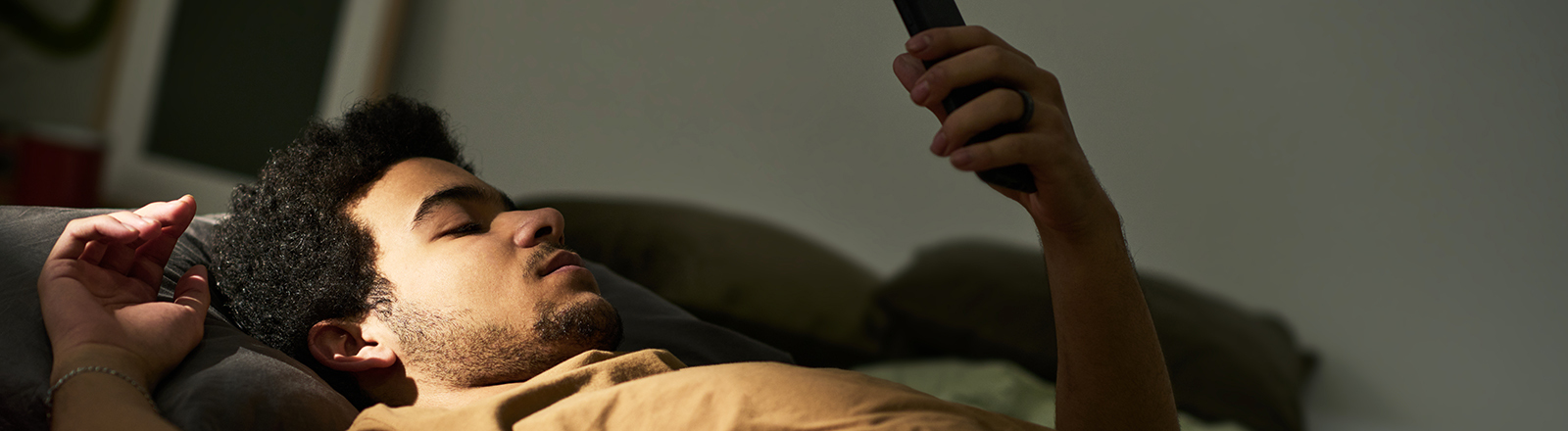 Ein Mann liegt auf dem Rücken im Bett. Sein linker Arm ist aufgestützt und er hält ein Smartphone in die Höhe, auf das er blickt. Es wirkt ruhig und etwas traurig.