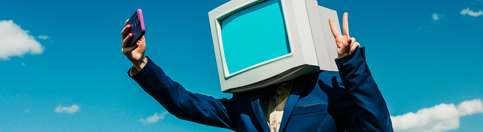 Eine Person in einem blauen Anzug trägt auf den Schultern einen Computerbildschirm, es ist eine Attrappe. Die Person macht ein Selfie von sich mit einem Smartphone.