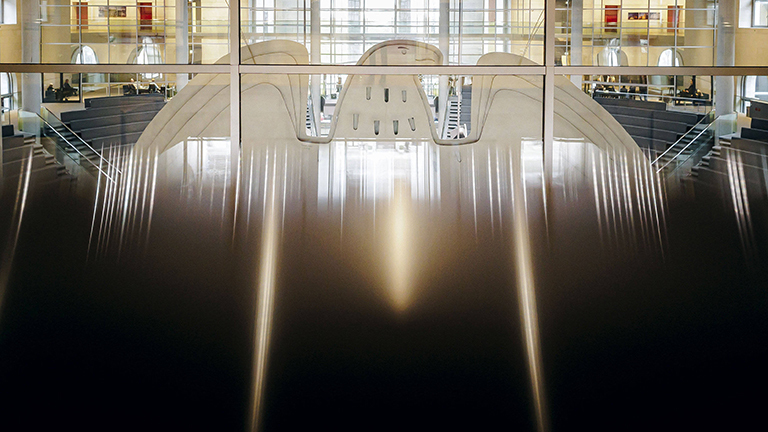Blick durch eine Glasfront von hinten auf den Bundesadler im Plenum des Bundestages.