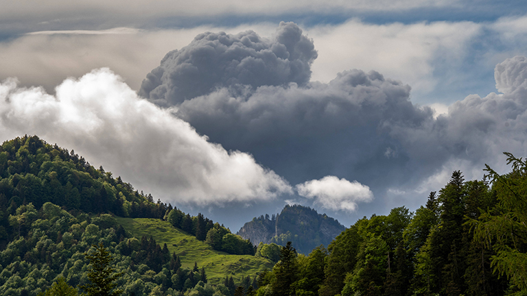 Hinter bewaldeten Bergen ziehen dunkle Gewitterwolken auf, es wirkt dramatisch.