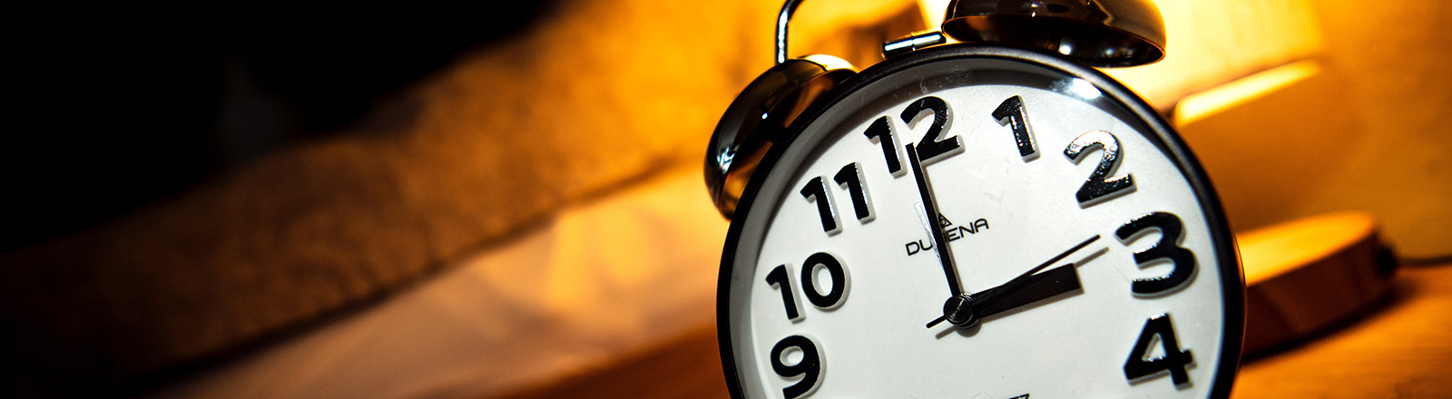 Ein Wecker steht neben einem Bett auf einem kleinen Tisch. Darauf steht auch eine Lampe, die warmes Licht macht. Der Wecker zeigt 3 Uhr.