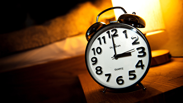 Ein Wecker steht neben einem Bett auf einem kleinen Tisch. Darauf steht auch eine Lampe, die warmes Licht macht. Der Wecker zeigt 3 Uhr.