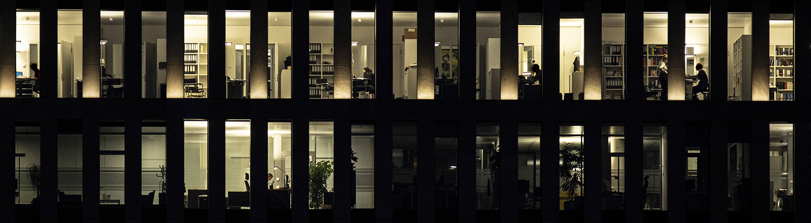 Beleuchtete Büroräume in der Dunkelheit. In wenigen Räumen sitzen Menschen an Schreibtischen.