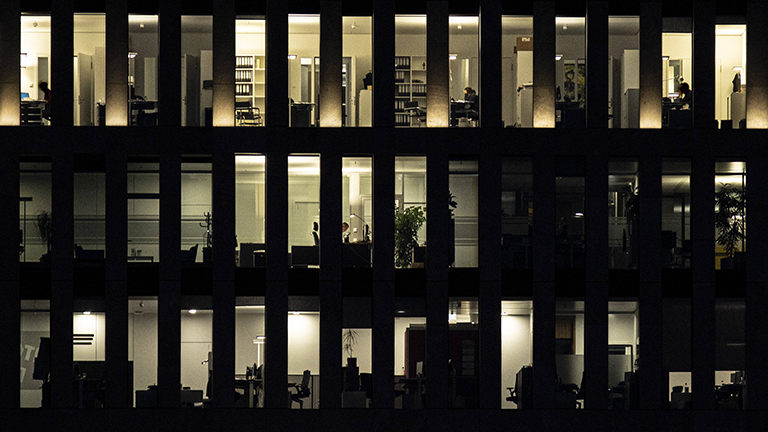 Beleuchtete Büroräume in der Dunkelheit. In wenigen Räumen sitzen Menschen an Schreibtischen.