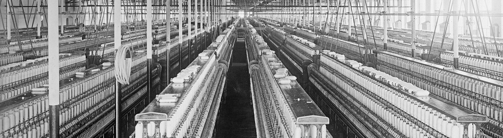 Schwarz-weiß-Foto: Blick in eine Baumwollspinnerei zwischen 1909 und 1920 in Manchester; eine große, menschenleere Fabrikhalle voller Webstühle.