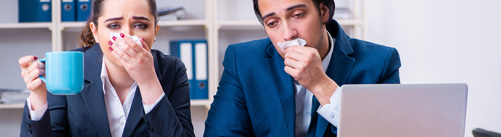 Symbolfoto: Ein Mann und eine Frau sitzen hinter einem Schreibtisch. Sie trägt einen blauen Blazer, er eine blaue Anzugsjacke. Sie halten sich Taschentücher vor den Mund und wirken sehr krank. Die Situation wirkt grotesk-lustig.