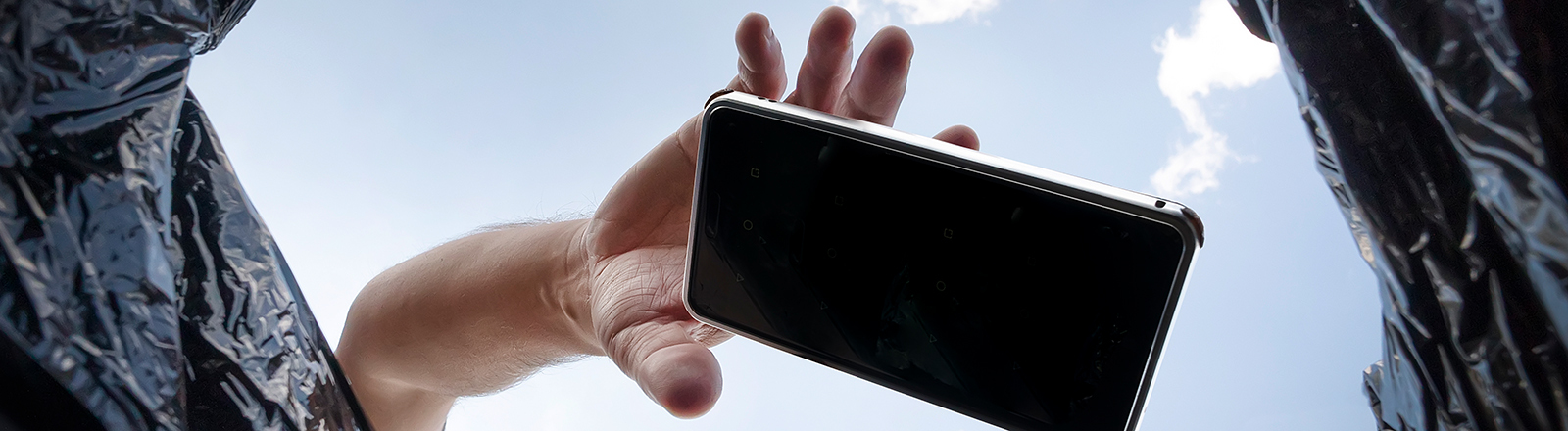 Blick aus einem Mülleimer in den blauen Himmel. Eine Hand wirft ein Smartphone in den Müll.
