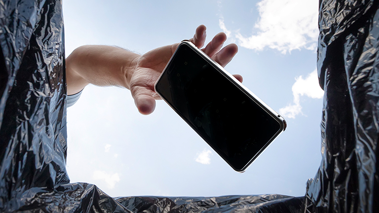 Blick aus einem Mülleimer in den blauen Himmel. Eine Hand wirft ein Smartphone in den Müll.