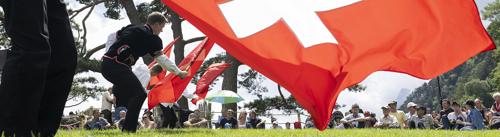 Auf der Rütliwiese in der Schweiz stehen Fahnenschwinger in traditioneller Kleidung; sie schwingen Schweizer-Flaggen (ein weißes Kreuz auf rotem Grund). Auf der Wiese sitzen Menschen und schauen zu. Es ist die Bundesfeier am 1. August 2025; Foto: dpa.