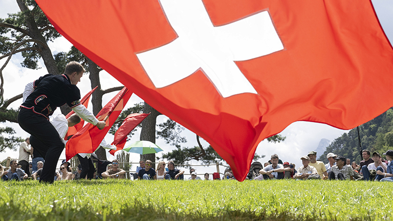 Auf der Rütliwiese in der Schweiz stehen Fahnenschwinger in traditioneller Kleidung; sie schwingen Schweizer-Flaggen (ein weißes Kreuz auf rotem Grund). Auf der Wiese sitzen Menschen und schauen zu. Es ist die Bundesfeier am 1. August 2025; Foto: dpa.