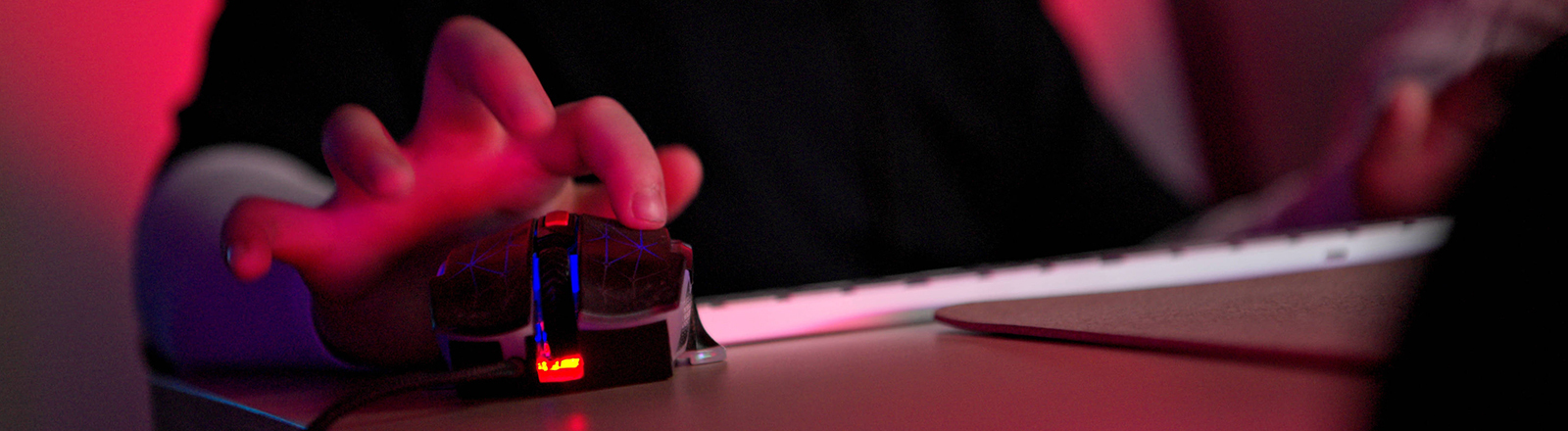 Nahaufnahme einer Person, die eine Gaming-Maus mit RGB-Beleuchtung neben einer Tastatur in einem dunklen Raum mit neonrotem Licht verwendet. Die Person ist nicht zu sehen, lediglich Oberkörper und die Hand auf der Gaming-Maus.