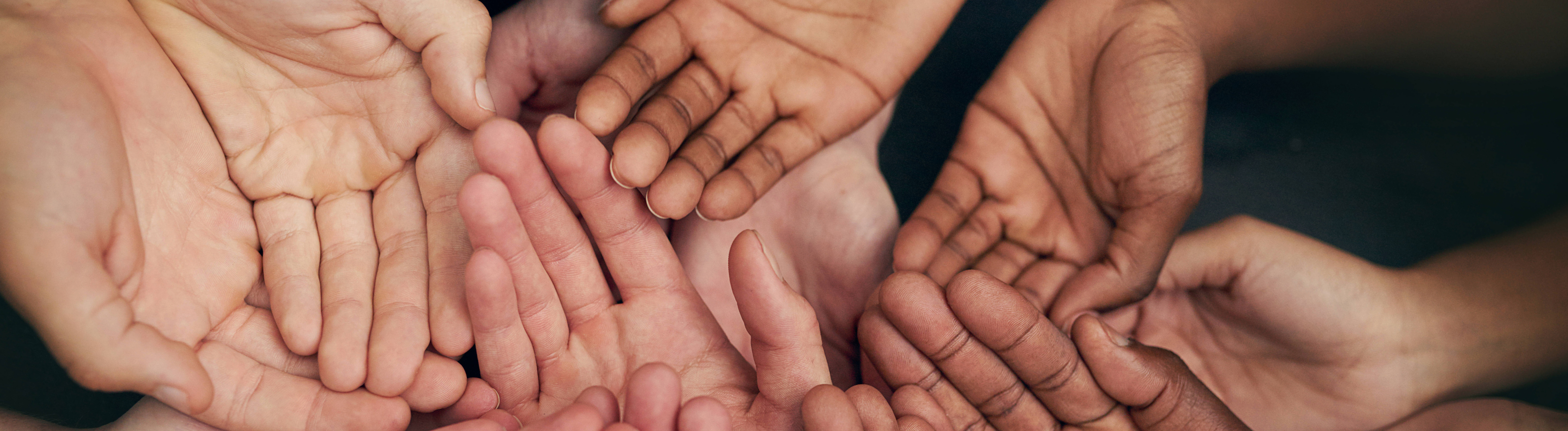 Viele Hände mit verschiedenen Hautfarben liegen übereinander, es wirkt nach Gemeinschaft.