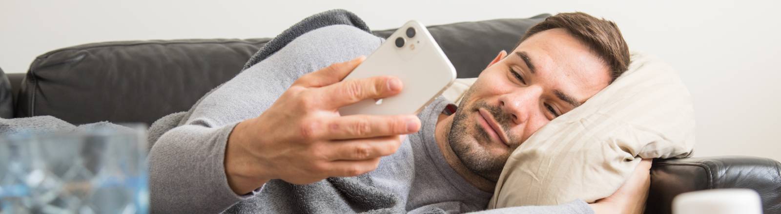 Ein Mann liegt auf einem Sofa und schaut aufs Smartphone, aufm dem Tisch vor ihm liegen u. a. Medikamente und ein Fieberthermometer