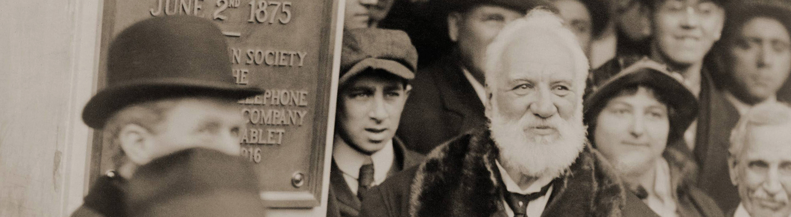 Schwarz-weiß Archifoto: Der Erfinder Graham Bell steht inmitten einer Menschengruppe. An der Wand neben ihm wurde ein Schild angebracht, das ihn als Erfinder des Telefons ehrt.