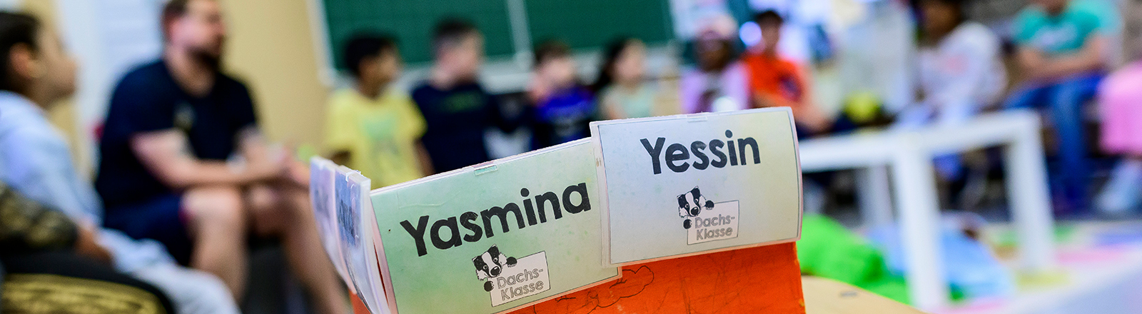 Schüler einer Grundschuleklasse sitzen vor der Tafel. Ebenso der Lehrer. Im Fotovordergrund steht auf einem Tisch eine Box mit zwei Namenskarten mit Yasmina und Yessin.