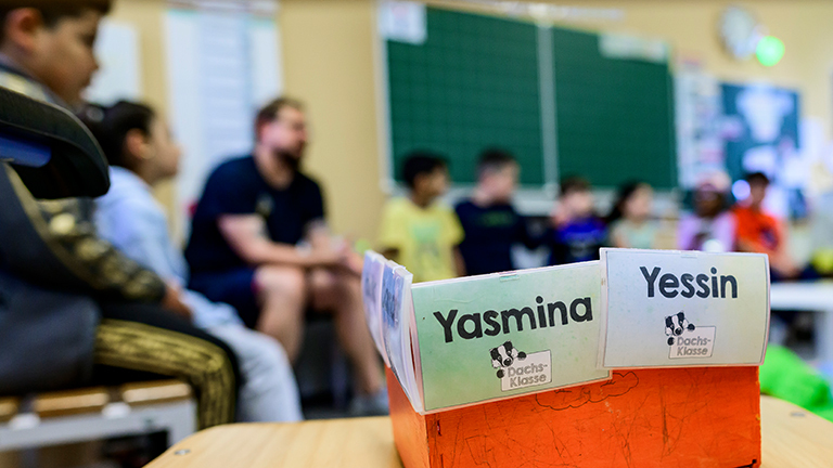 Schüler einer Grundschuleklasse sitzen vor der Tafel. Ebenso der Lehrer. Im Fotovordergrund steht auf einem Tisch eine Box mit zwei Namenskarten mit Yasmina und Yessin.