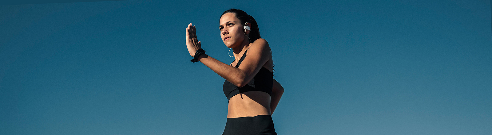 Eine junge Frau joggt draußen vor blauem Himmel. Sie wirkt sehr fit und dynamisch. Im Hintergrund sind Schornsteine zu sehen.