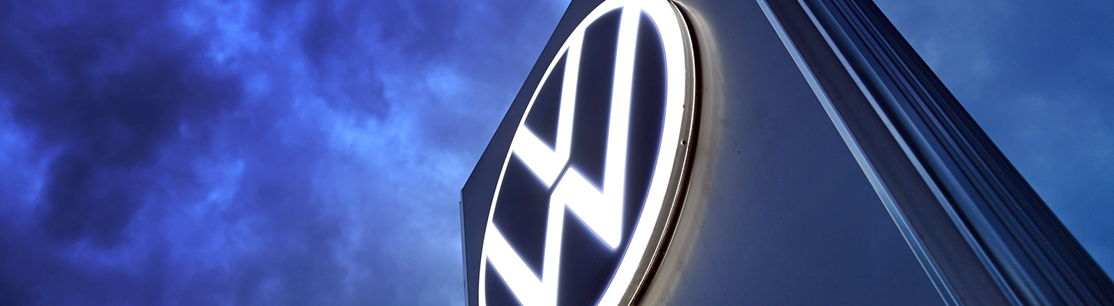 Das Volkswagen-Logo auf einer Steele vor dunklem Himmel mit vielen Wolken.