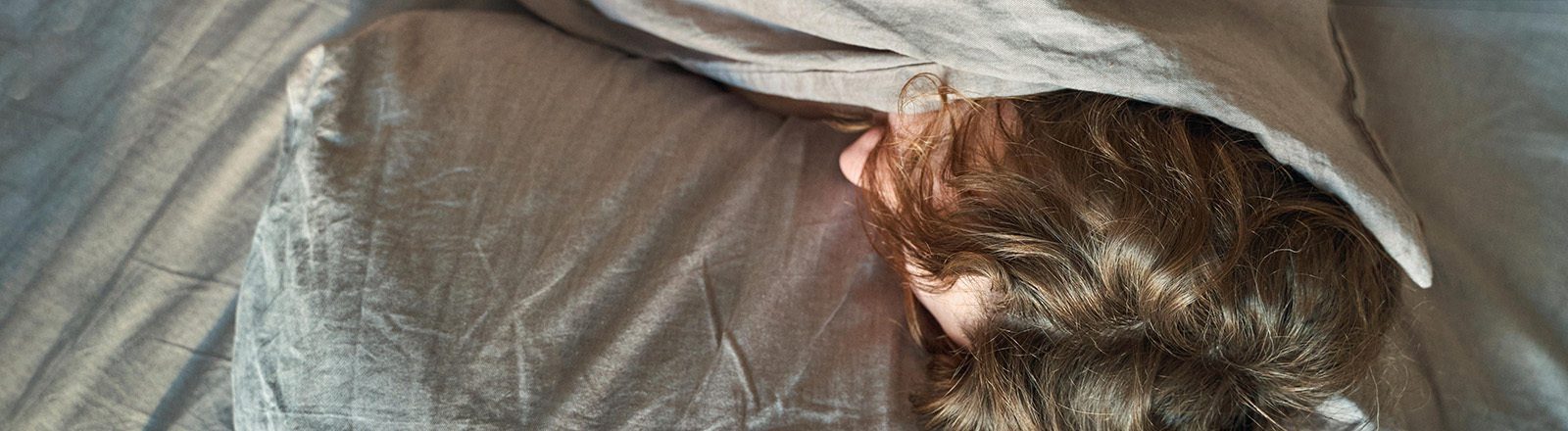 Eine Person liegt im Bett und hat die Bettdecke nach oben gezogen. Lediglich die etwas lockigen Haaren sind zu sehen.