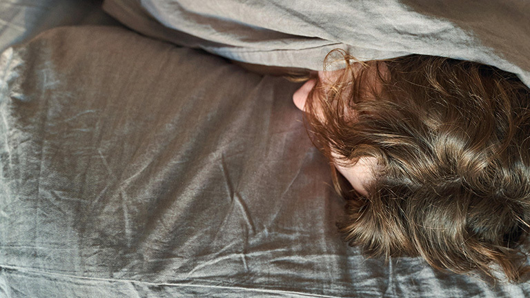 Eine Person liegt im Bett und hat die Bettdecke nach oben gezogen. Lediglich die etwas lockigen Haaren sind zu sehen.