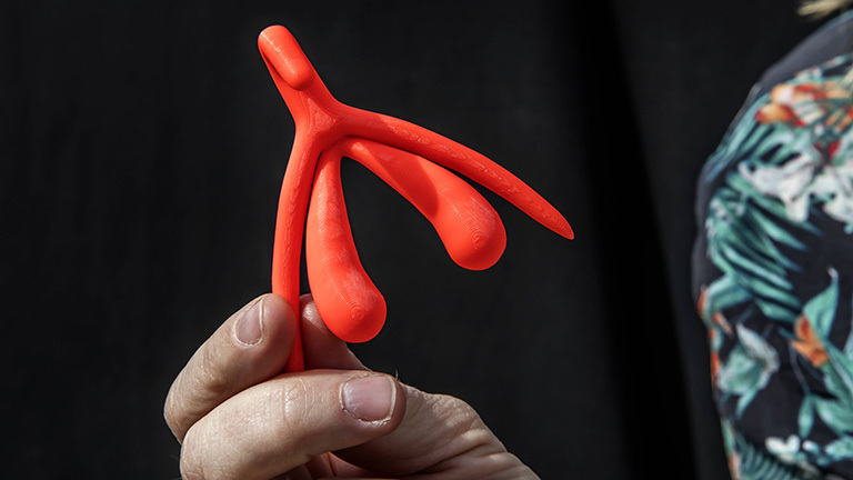Eine Hand hält einen 3D-Druck einer Klitoris in roter Farbe.