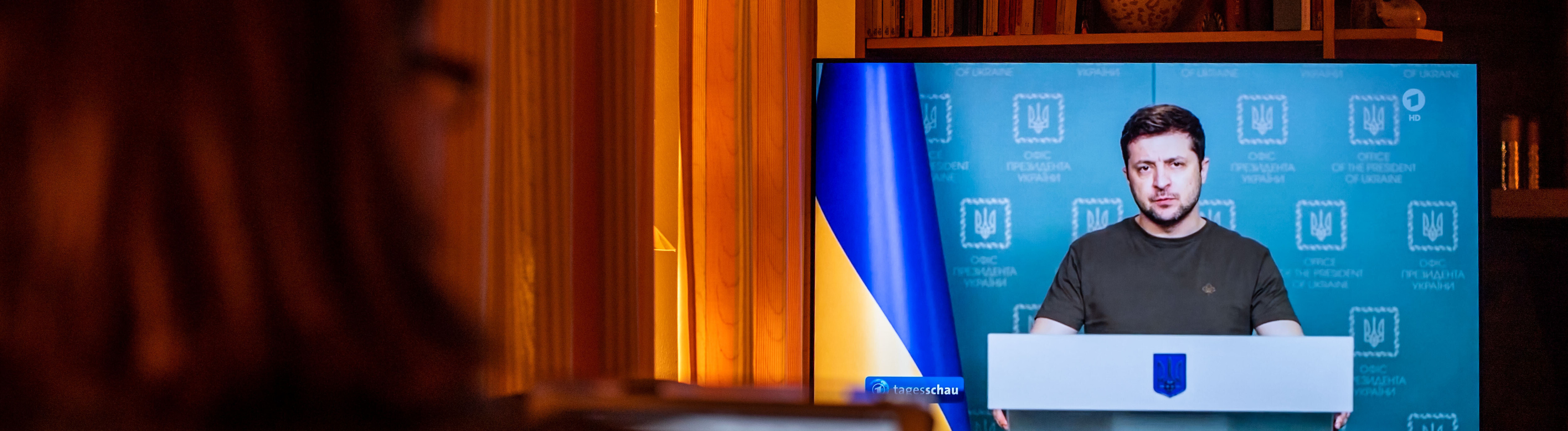 Eine Frau sitzt auf dem Sofa und sieht sich im Fernsehen die Nachrichtensendung "Tagesschau" an. Dort im Bild ist der ukrainische Präsident ukrainische Präsident Selenskyj. Die Aufnahme ist von März 2022.