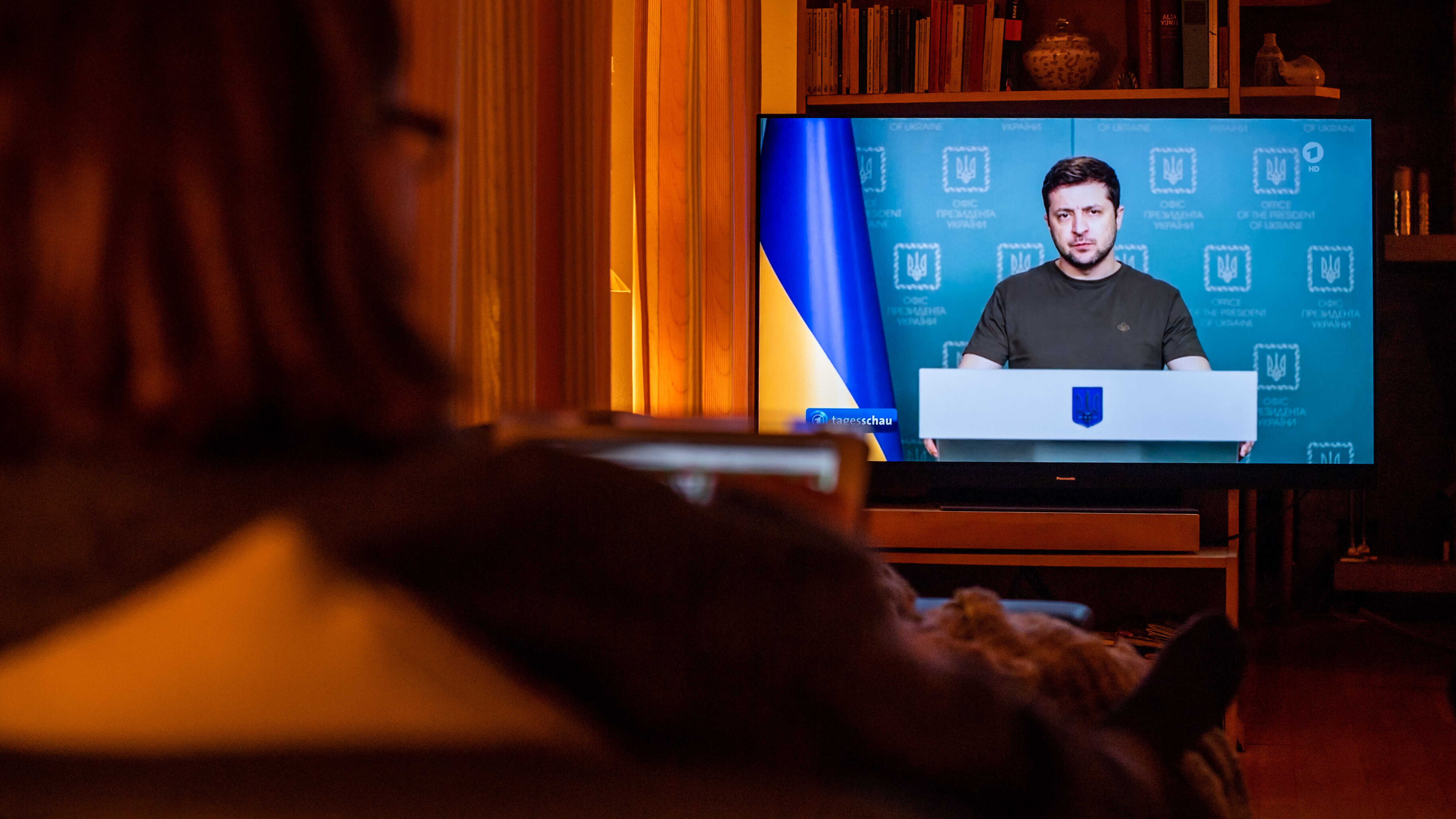 Eine Frau sitzt auf dem Sofa und sieht sich im Fernsehen die Nachrichtensendung "Tagesschau" an. Dort im Bild ist der ukrainische Präsident ukrainische Präsident Selenskyj. Die Aufnahme ist von März 2022.