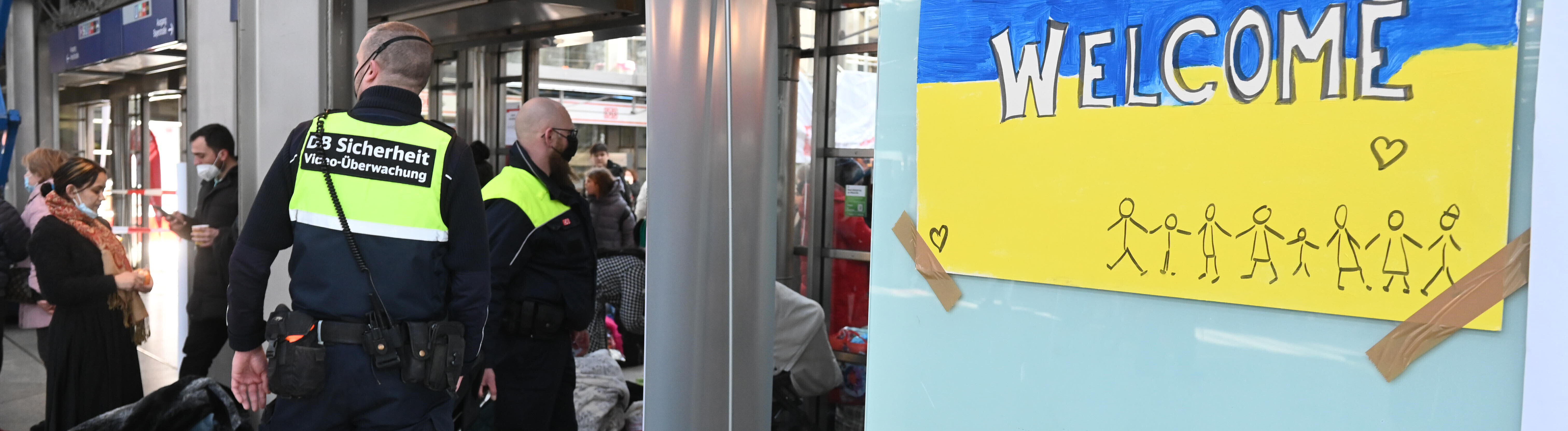 Mitarbeiter der DB-Sicherheit steht am Münchner Hauptbahnhof neben einem selbstgemalten Bild mit ukrainischer Flagge, der Aufschrift „welcome“ sowie Strichmännchen und einem Herzen (11.03.2022)