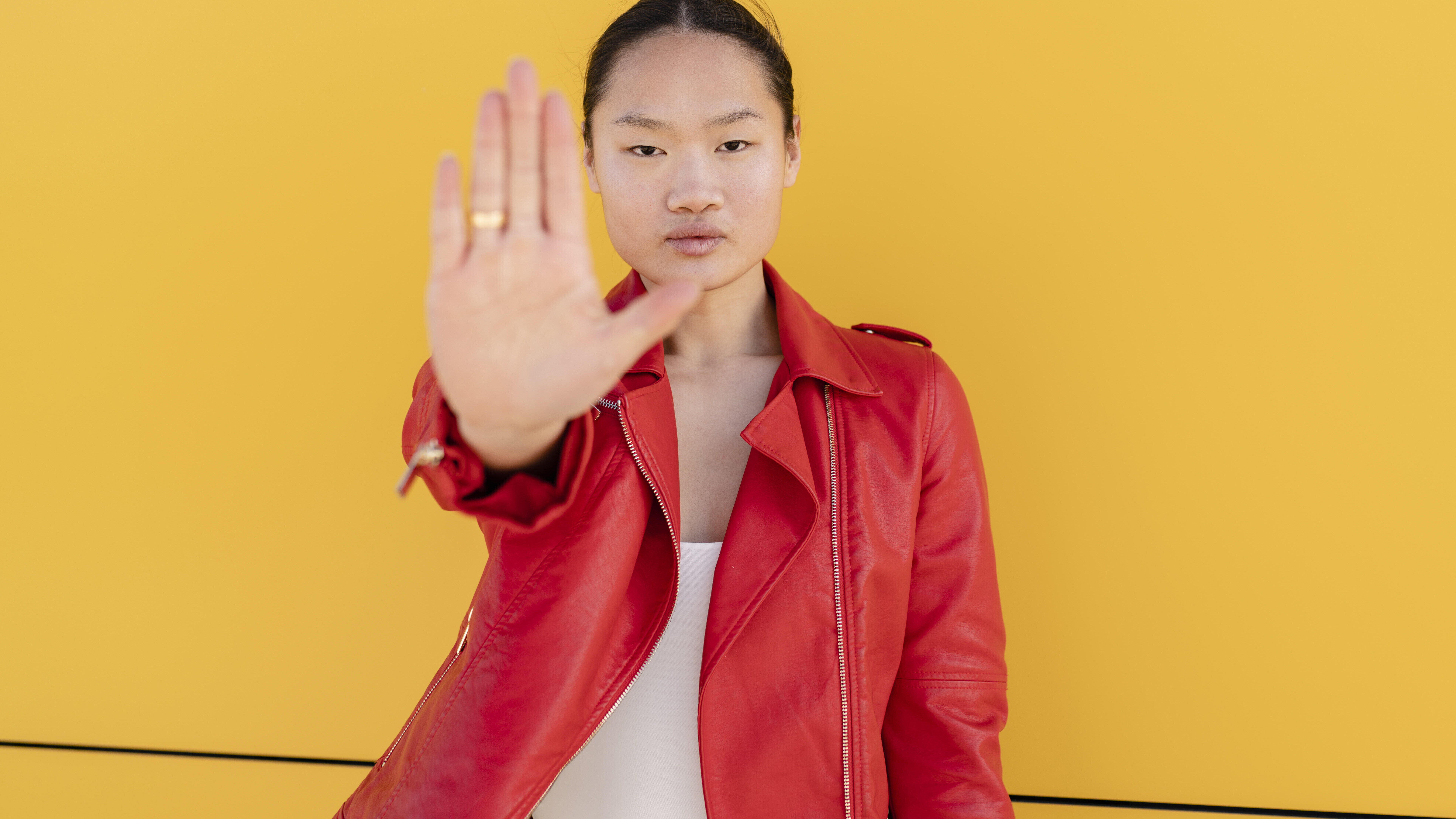 Eine Frau steht vor einer gelben Wand. Sie trägt eine rote Lederjacke, schaut in die Kamera und hält ihre flache Hand hoch zu einer Stopp-Geste.