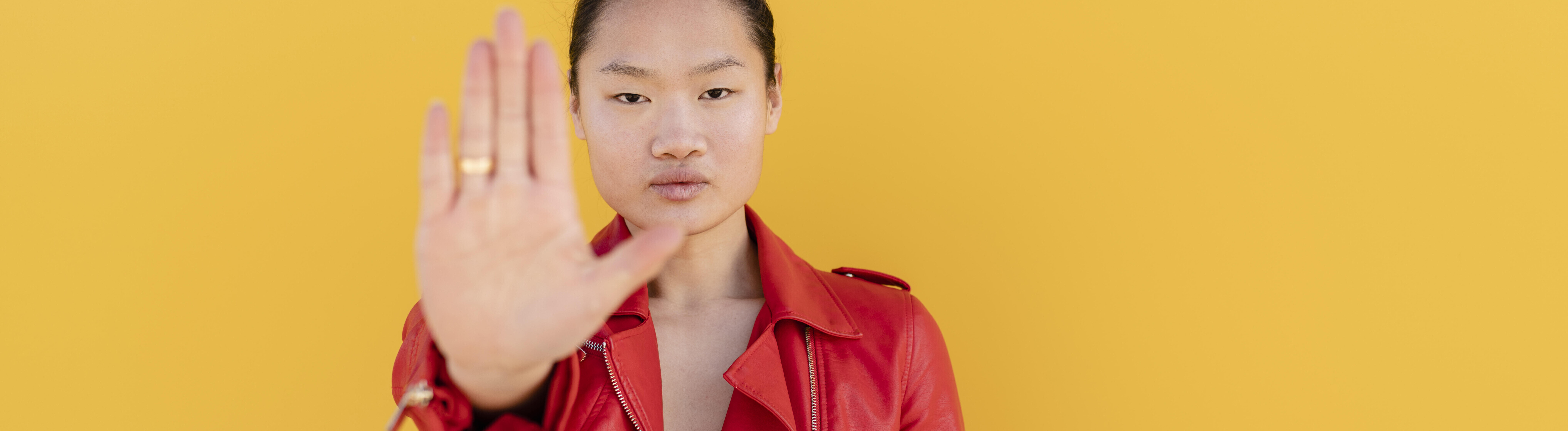 Eine Frau steht vor einer gelben Wand. Sie trägt eine rote Lederjacke, schaut in die Kamera und hält ihre flache Hand hoch zu einer Stopp-Geste.