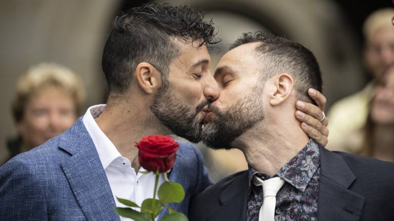 Zwei Männer küssen sich und halten eine rote Rose dabei. Sie tragen Anzüge und hinter ihnen stehen lächelnde Menschen.