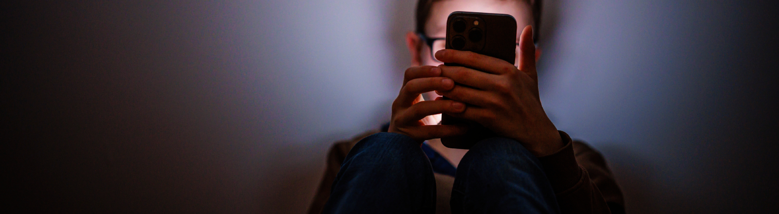 Ein Junge sitzt in einem dunklen Raum und wird von einem Smartphone beleuchtet.