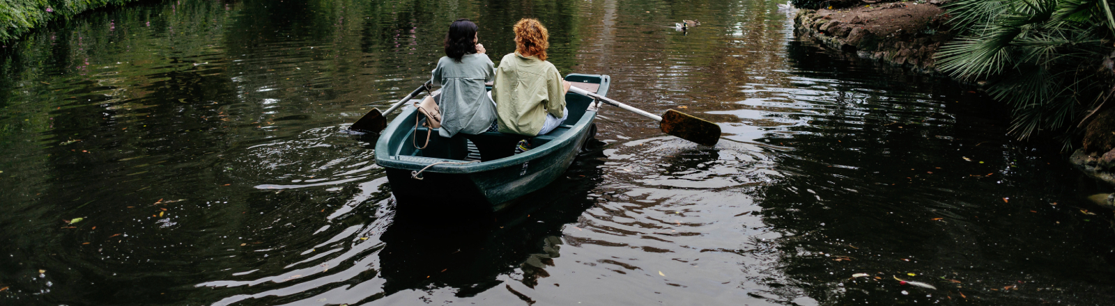 Lesbisches Pärchen in einem Ruderboot auf einem Weiher in Barcelona. Ringsum viel Grün.