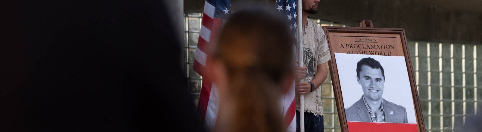 Ein Schwarz-Weiß-Foto on Charlie Kirk bei einer Trauerfeier, daneben die US-Flagge, im Vordergrund Menschen