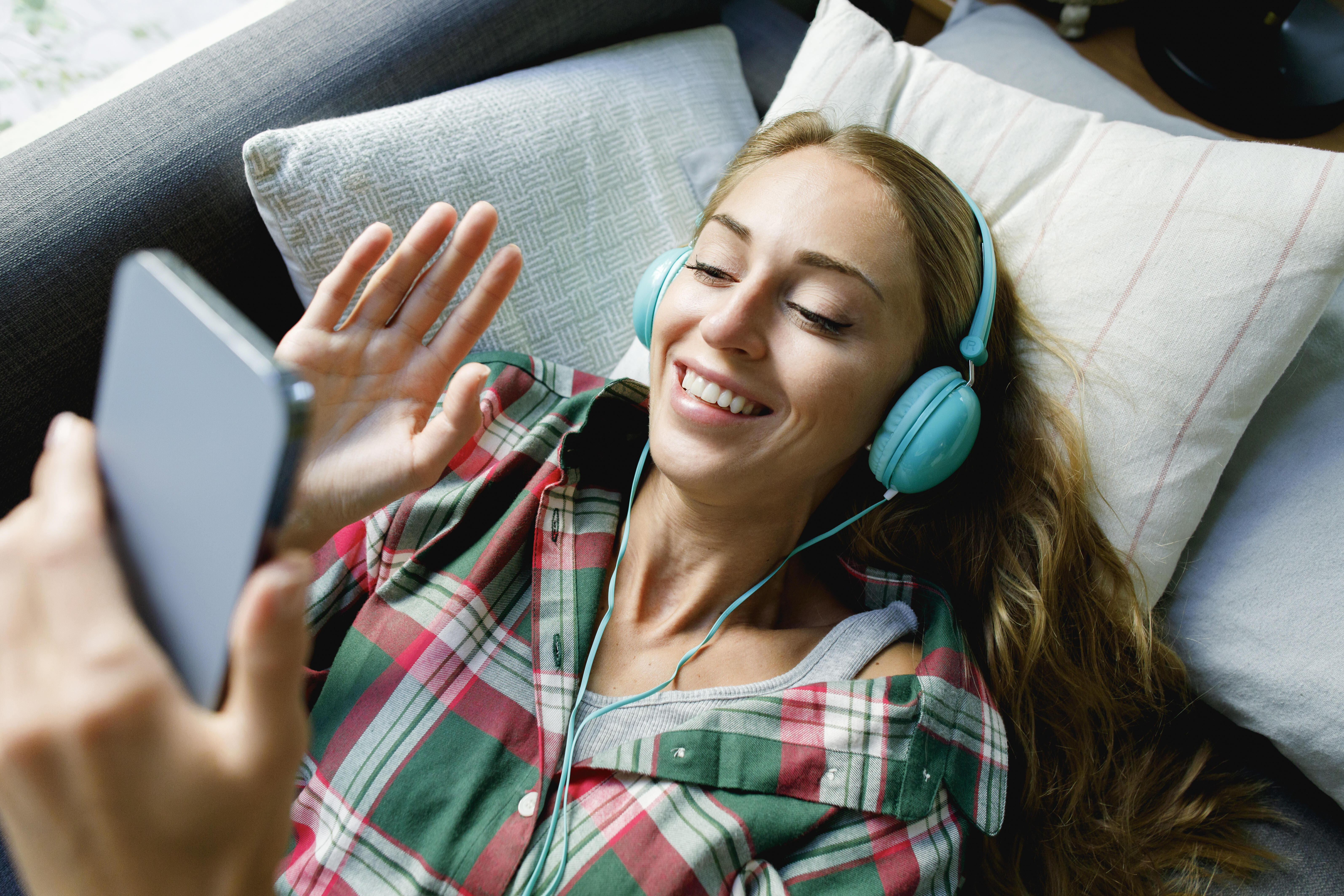 Frau liegt auf Sofa, sie trägt Kopfhörer und winkt lächelnd ins Smartphone