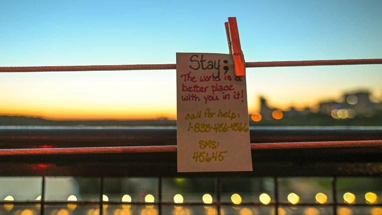 An einer Brückenvertstrebung hängt ein Zettel mit der Aufschrift: „Stay: The World is a better place with you in it.“ Im Hintergrund sieht man die Lichter einer Stadt.