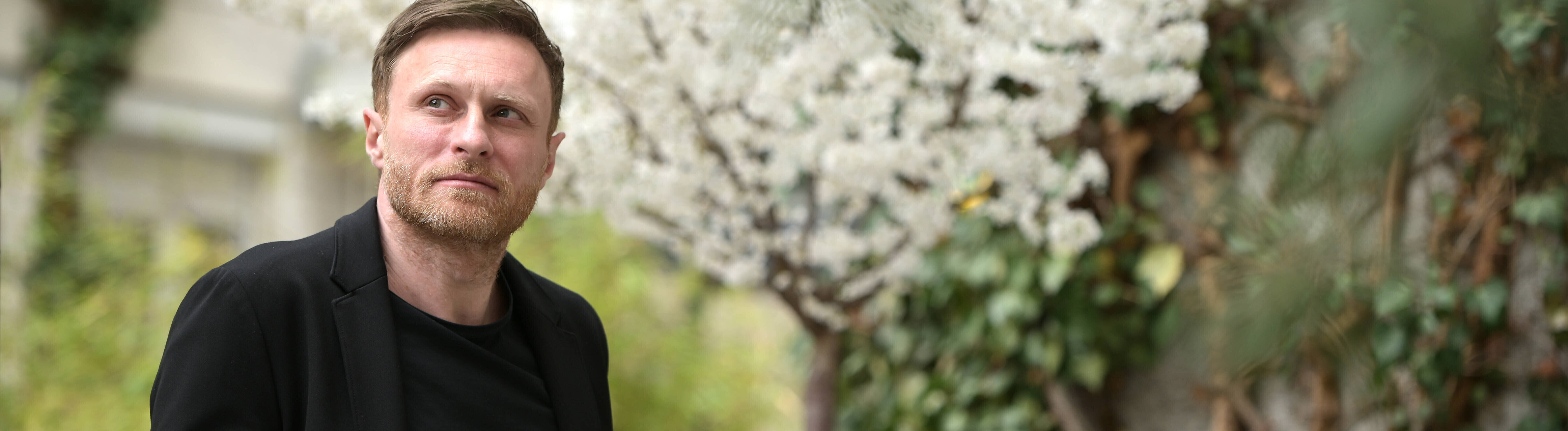 Der deutsche Autor Michael Nast steht draußen in einem Garten, er trägt ein schwarzes Jackett und Oberteil. Im Hintergrund blüht ein weißer Kirschbaum.